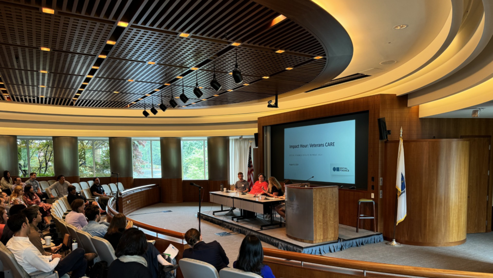 A panel of three people sits at a table in a conference room, presenting to an audience seated in rows. A screen displays the text "Impact Year: Veterans CARE.